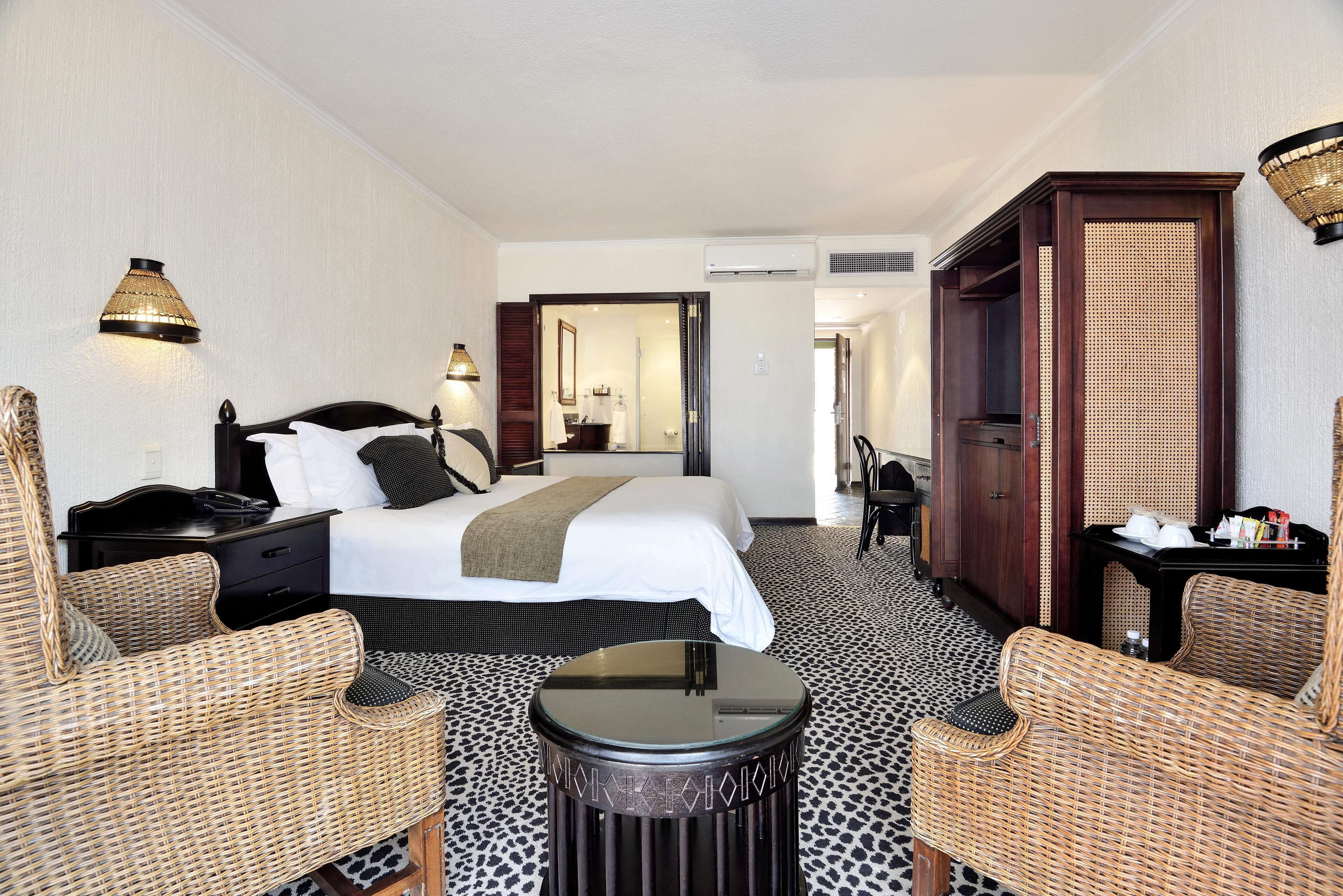A hotel room with a bed, two wicker chairs, a small table, a wardrobe, and a mirror.