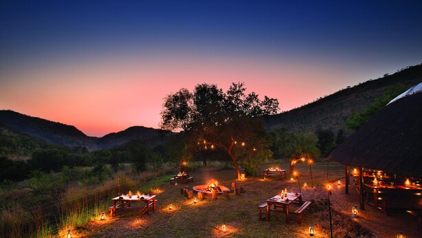 An outdoor dining area with tables and chairs arranged around a central fire pit, surrounded by lanterns and a thatched-roof structure. - Bakubung Bush Lodge (Pilanesberg National Park)