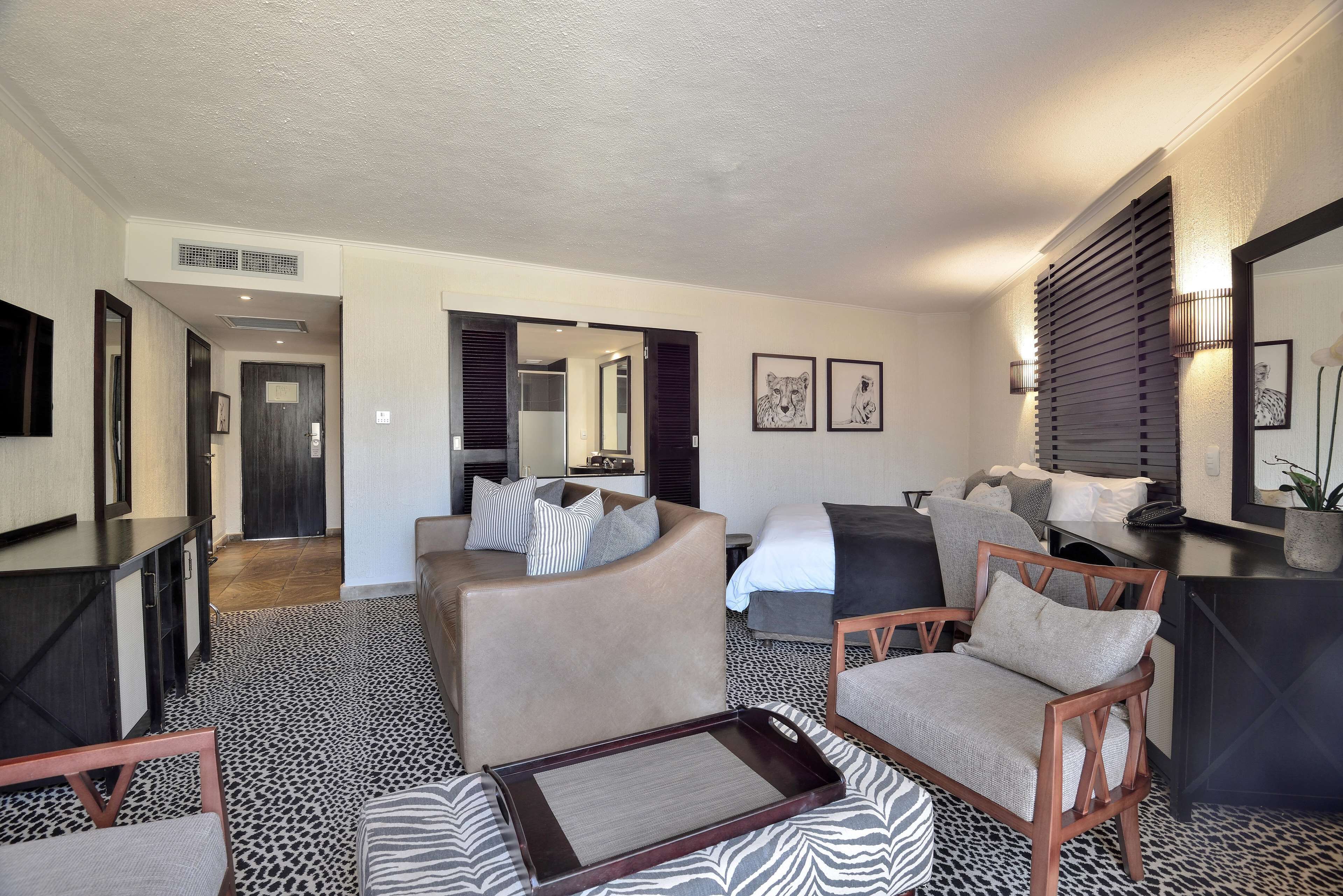 A hotel room with a sofa, armchair, and a tray table. There is a bed with a headboard and a bedside table. The room has a patterned carpet and a large mirror on the wall.