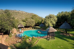 Una piscina con sillas de descanso y sombrillas de paja, rodeada de vegetación y un paisaje montañoso.