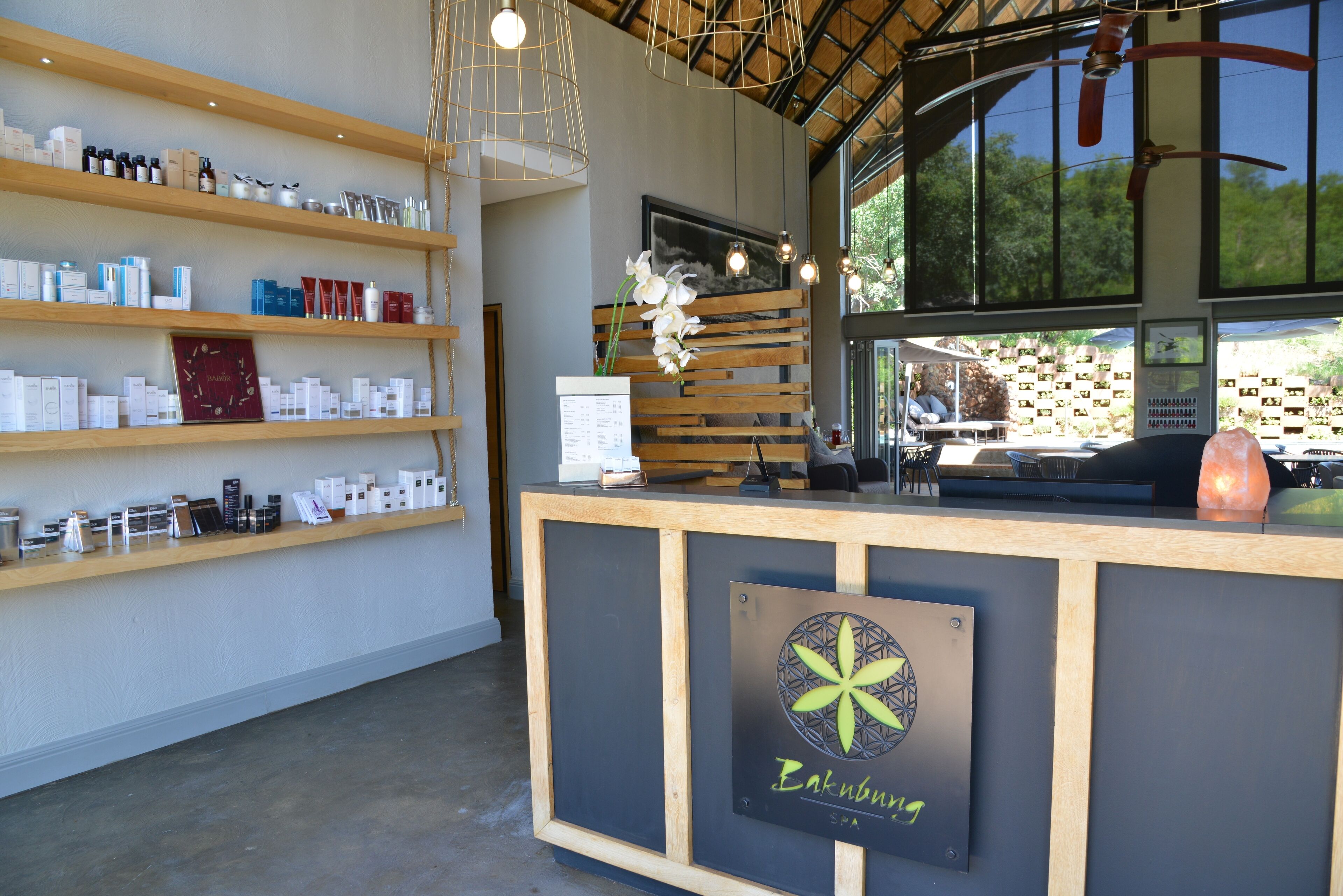 A reception desk with a sign reading "Bakmbury," shelves with various products, and a large window with a view of greenery.