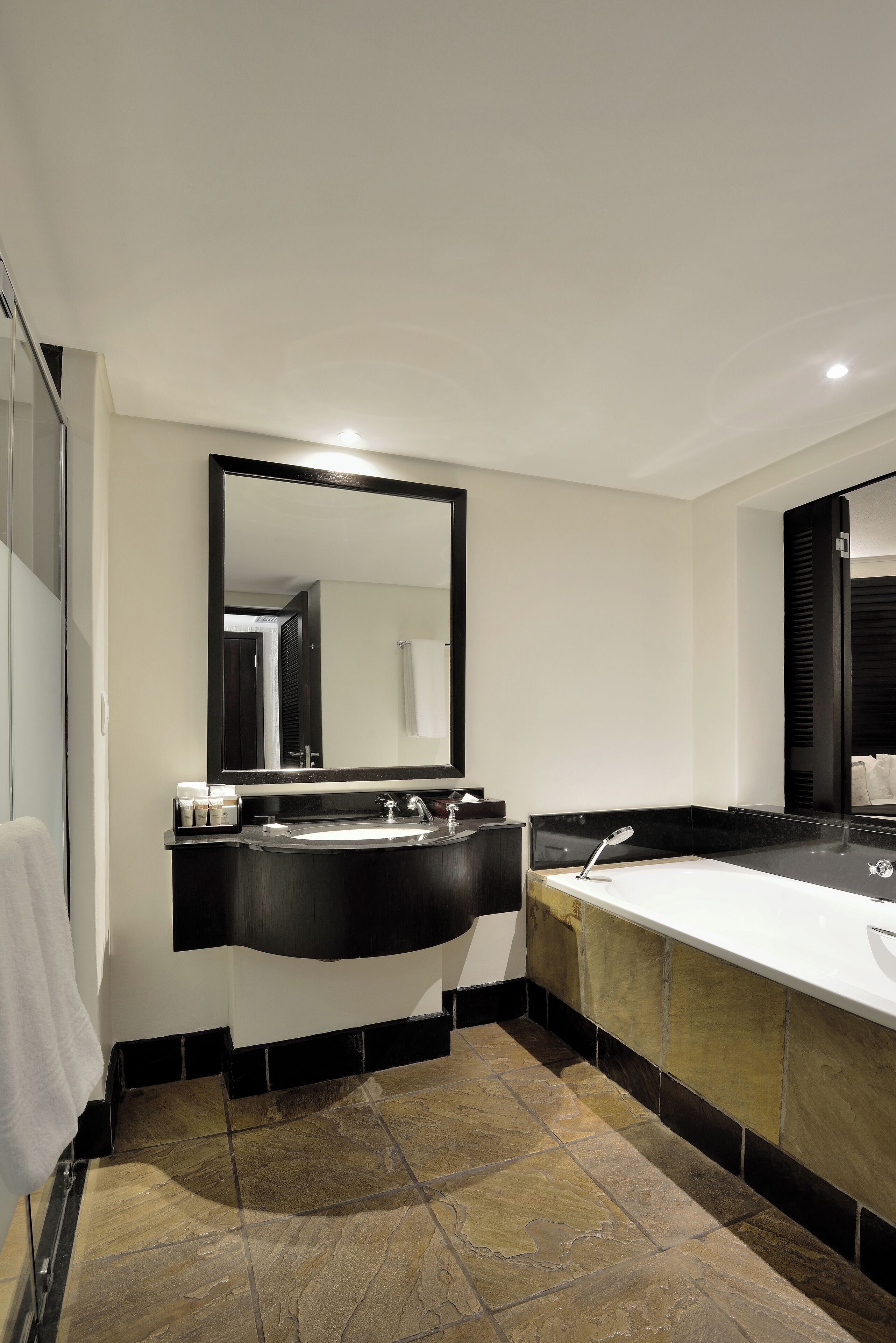 A modern bathroom with a black vanity, a large mirror, a white bathtub, and stone tiles.