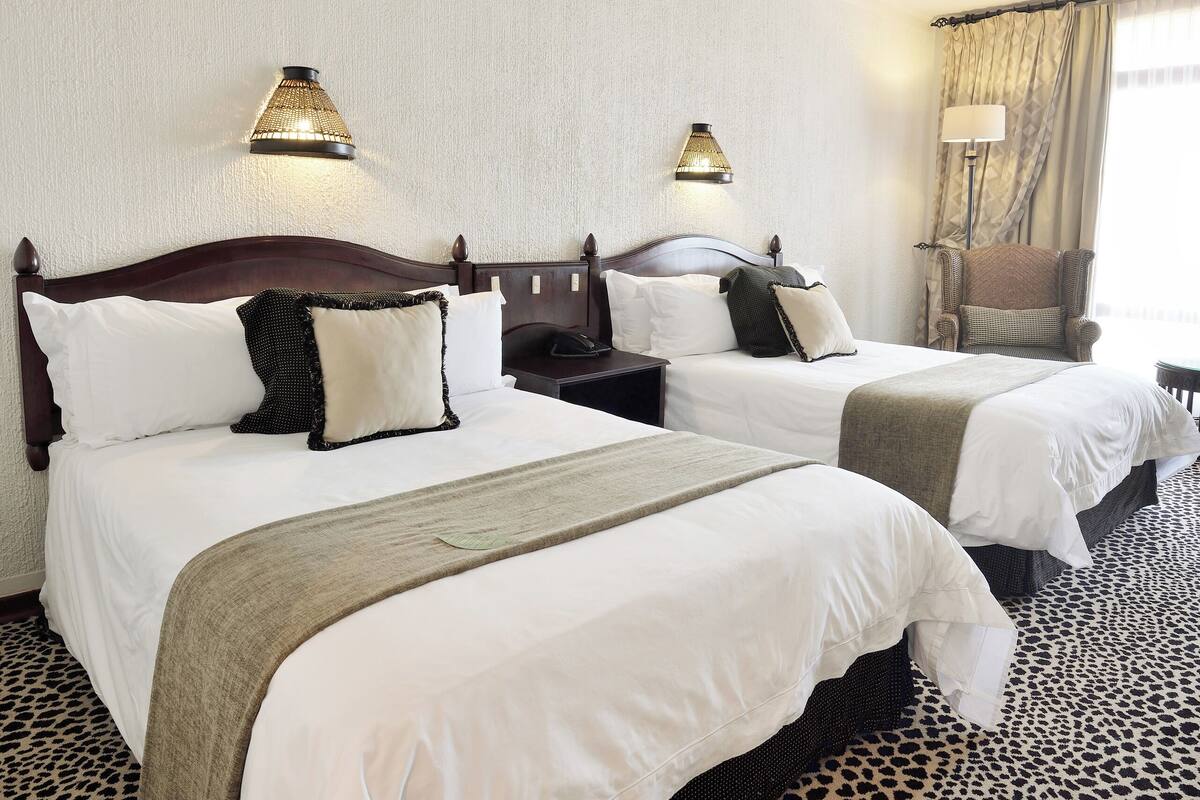 Habitación de hotel con dos camas, cada una con ropa blanca, cabeceros oscuros y una lámpara en la mesita de noche. La habitación tiene una alfombra de estampado de leopardo y una ventana con cortinas.