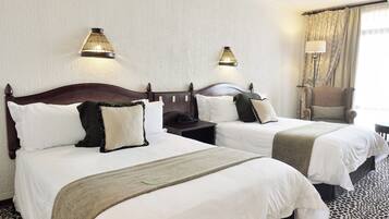 Quarto de hotel com duas camas, cada uma com roupa de cama branca, cabeceira escura e abajur na mesa de cabeceira. O quarto possui um tapete com estampa de leopardo e uma janela com cortinas.