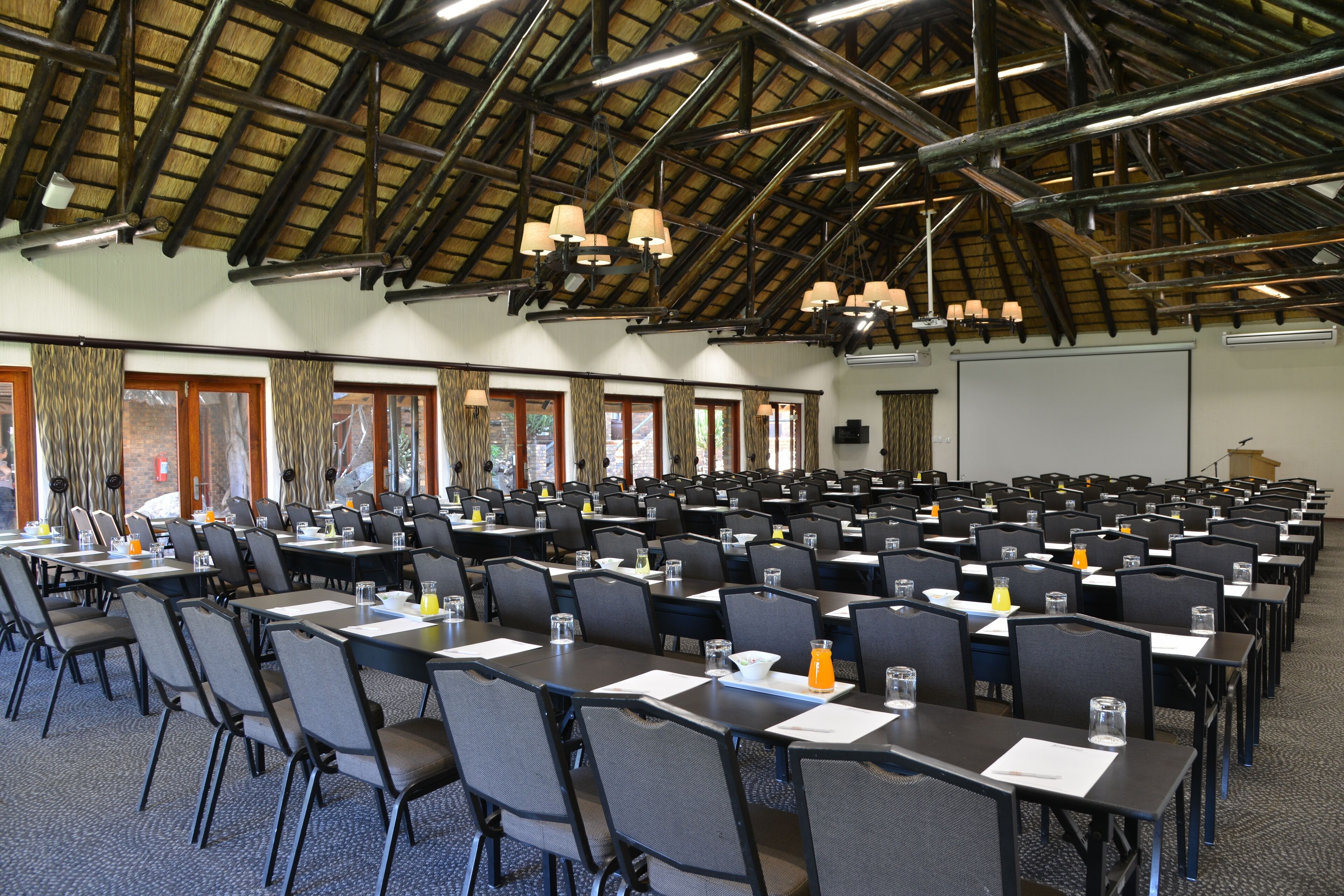 Un salón de conferencias con filas de mesas y sillas, un techo alto con vigas de madera y amplias ventanas.
