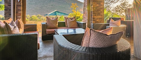 An outdoor patio with wicker furniture, a glass table, and a view of a hilly landscape.