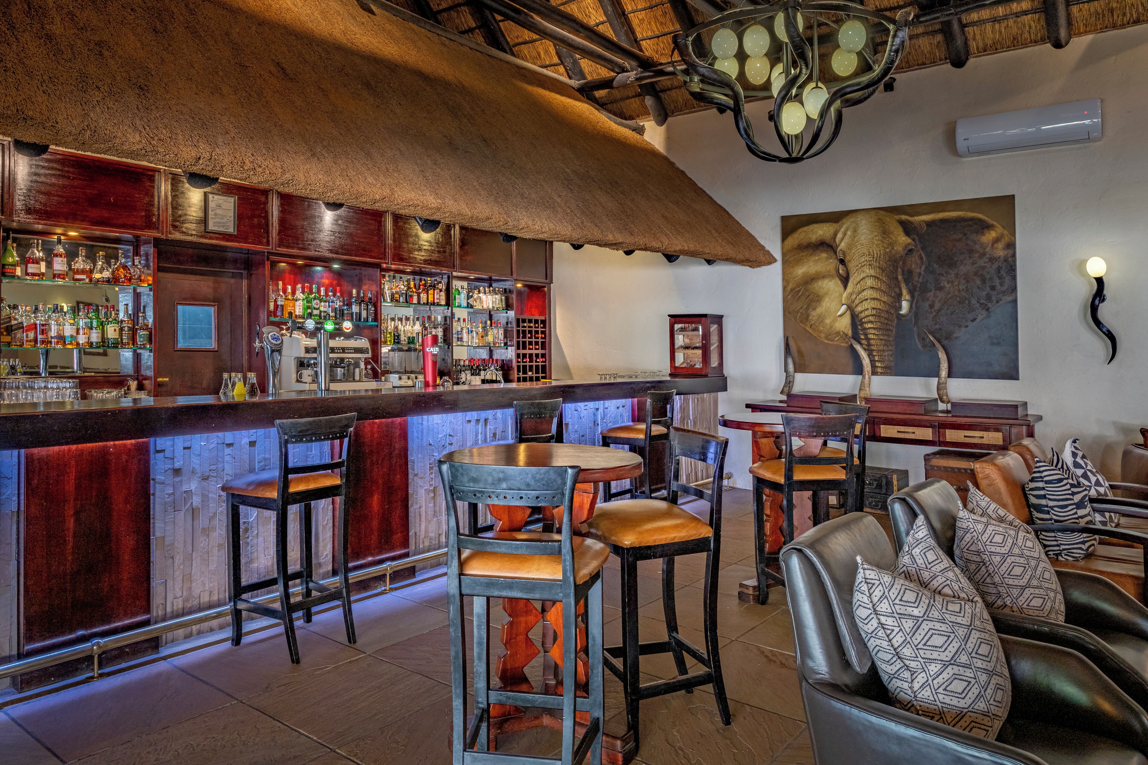 A bar area with wooden bar counter, stools, and a large elephant painting on the wall.