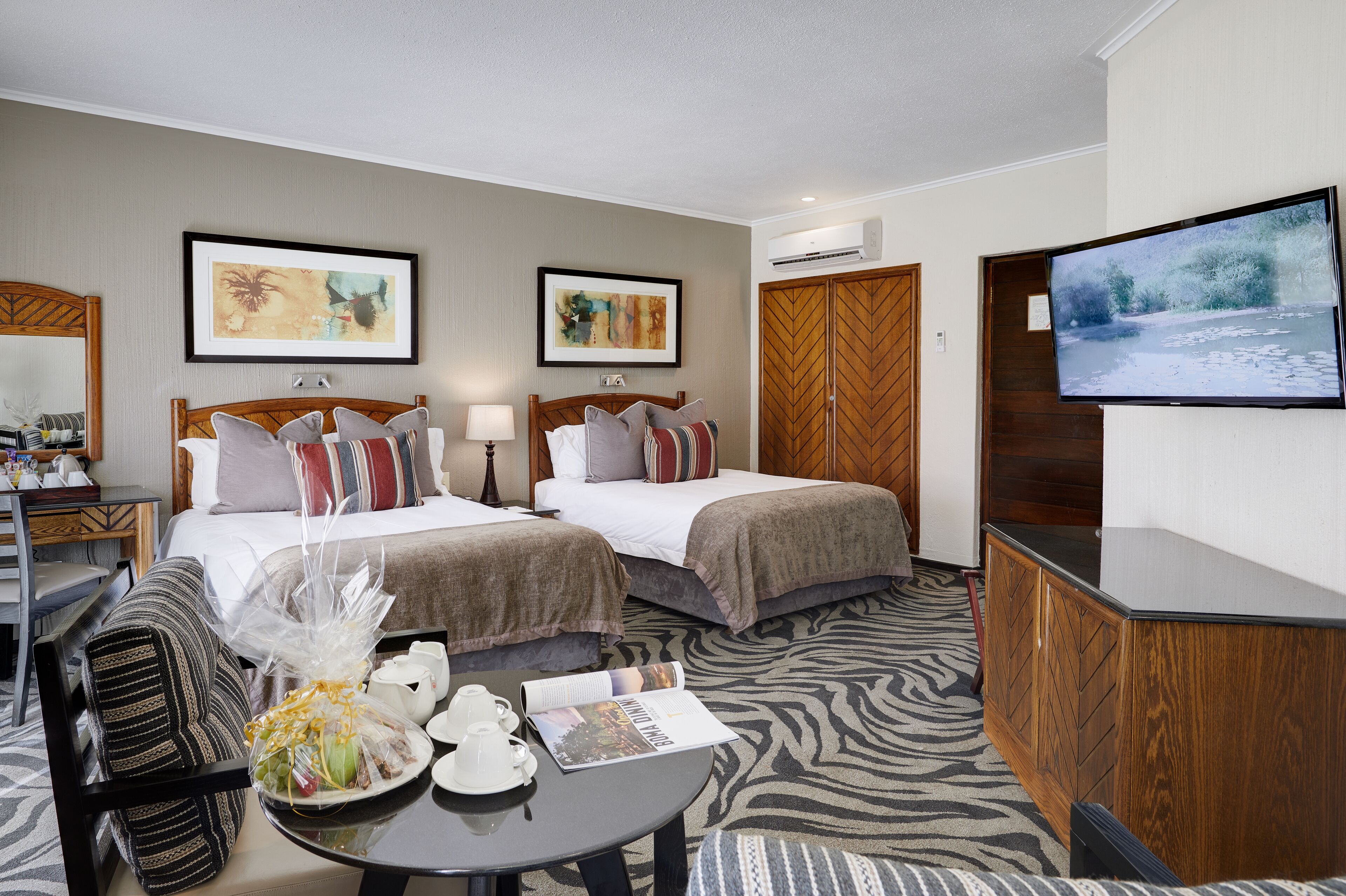 A hotel room with two beds, a TV, a desk with a lamp, and artwork on the walls.