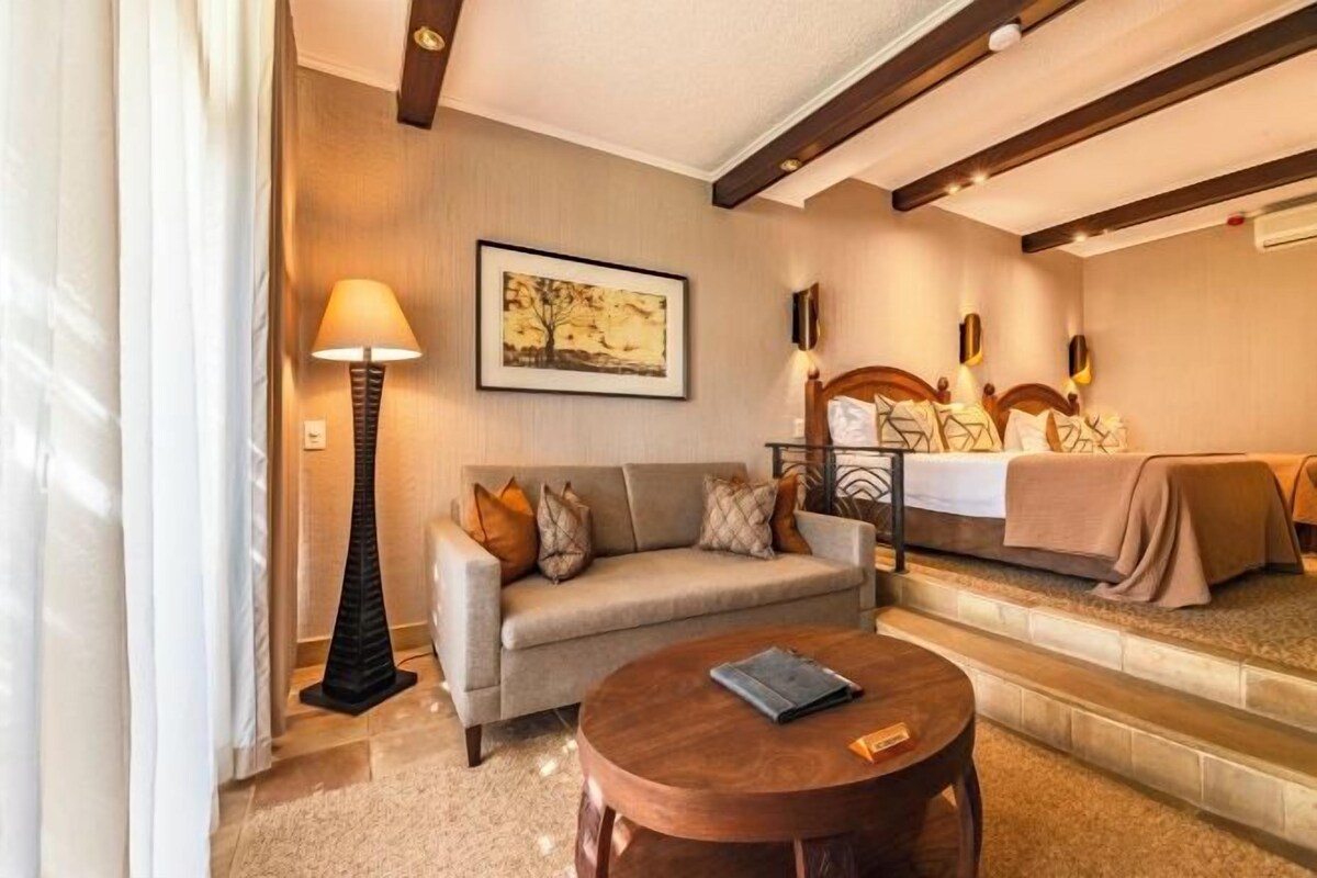 Habitación de hotel con sofá, una mesa redonda de madera, lámpara, cama con cabecera y un cuadro en la pared.