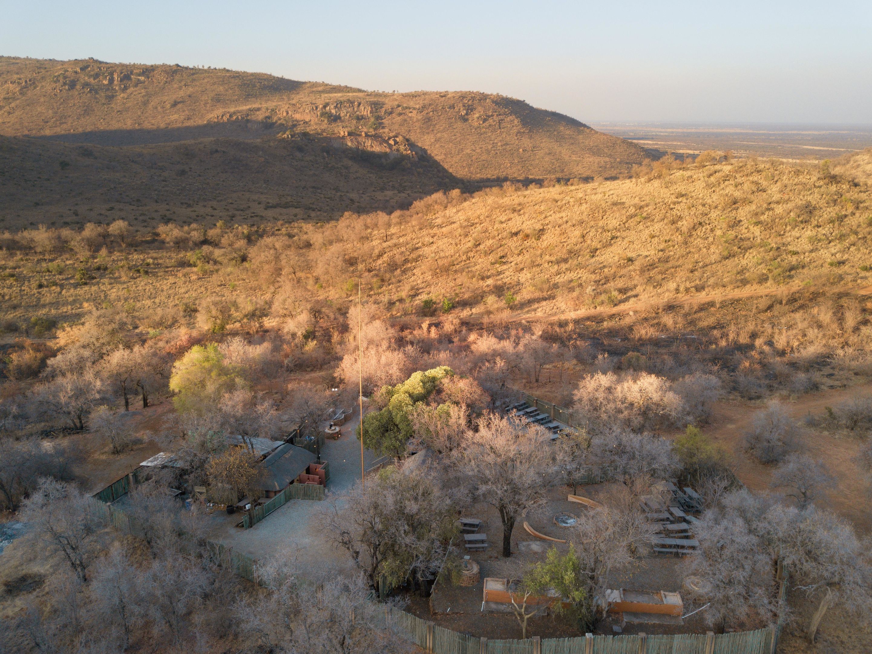 A rural landscape with a fenced area containing several wooden structures, possibly cabins or sheds, surrounded by dry vegetation and hills in the background.