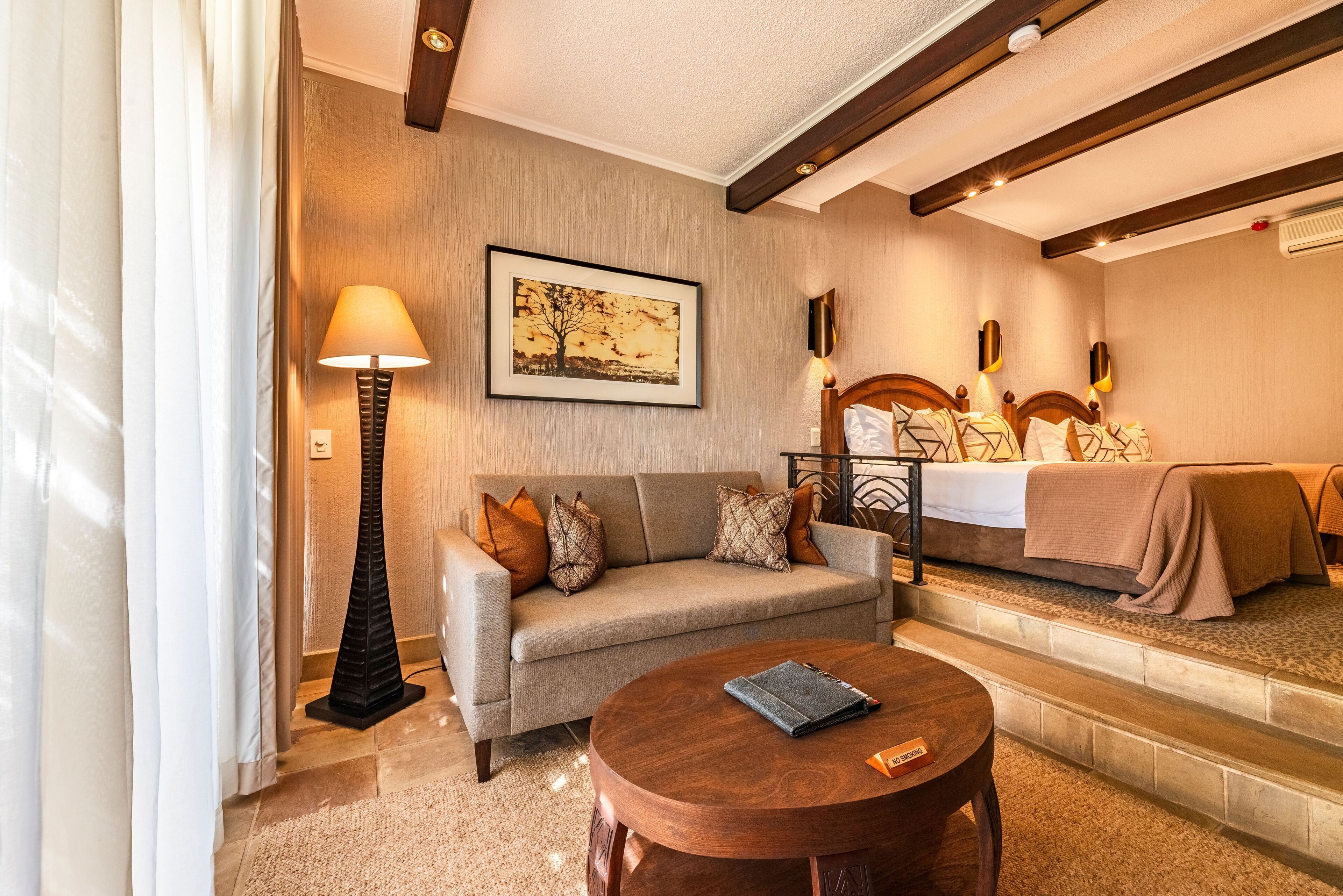 A hotel room with a sofa, a round wooden table, a bed with a headboard, a floor lamp, and a framed picture on the wall.