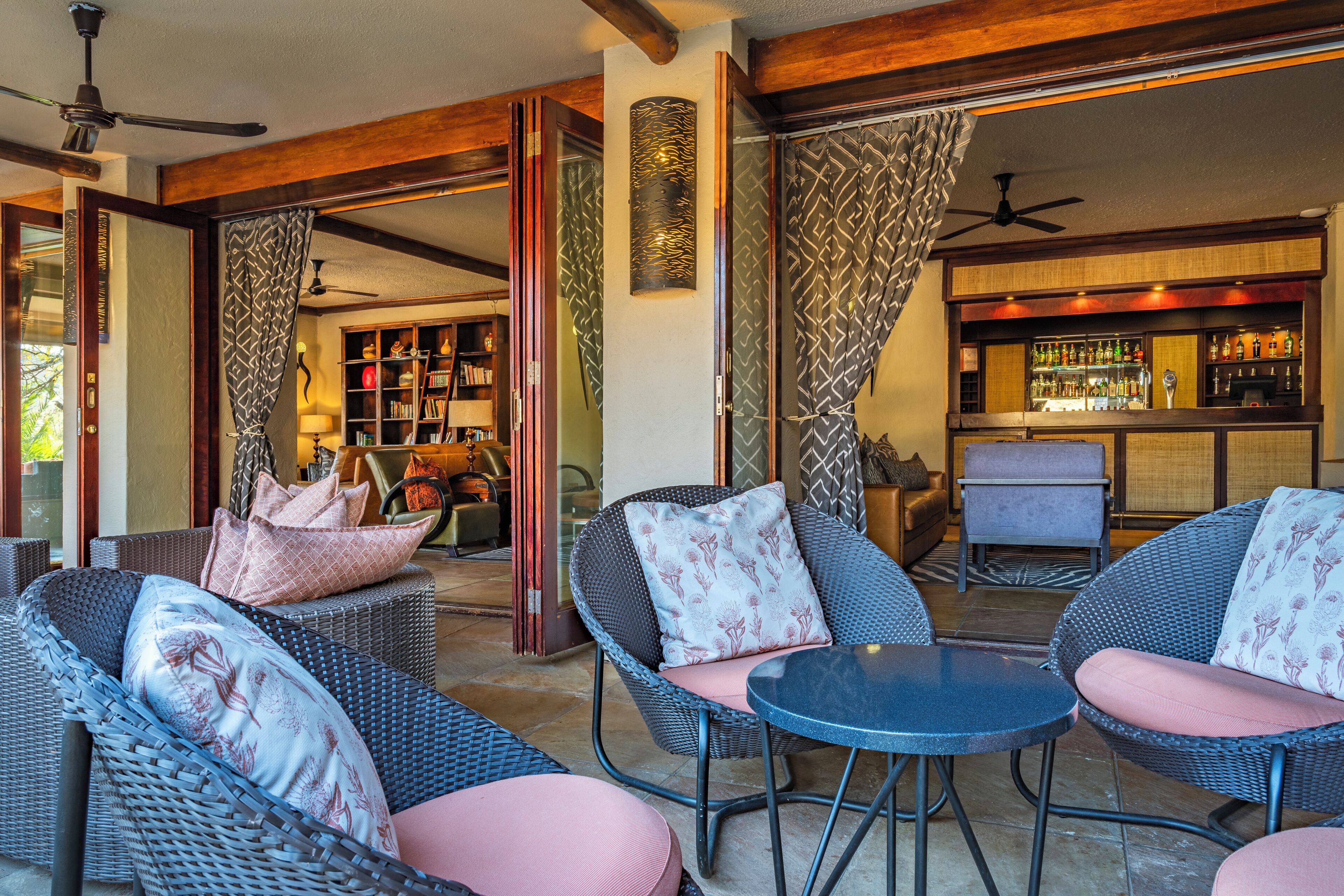 A cozy outdoor seating area with wicker furniture and cushioned chairs, a bar with a variety of bottles, and a bookshelf filled with books.