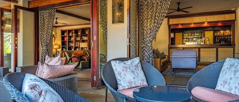 A cozy outdoor seating area with wicker furniture and cushioned chairs, a bar with a variety of bottles, and a bookshelf filled with books.