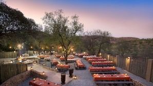An outdoor dining area with multiple tables set for guests, surrounded by a wooden fence, and featuring a fire pit in the center.