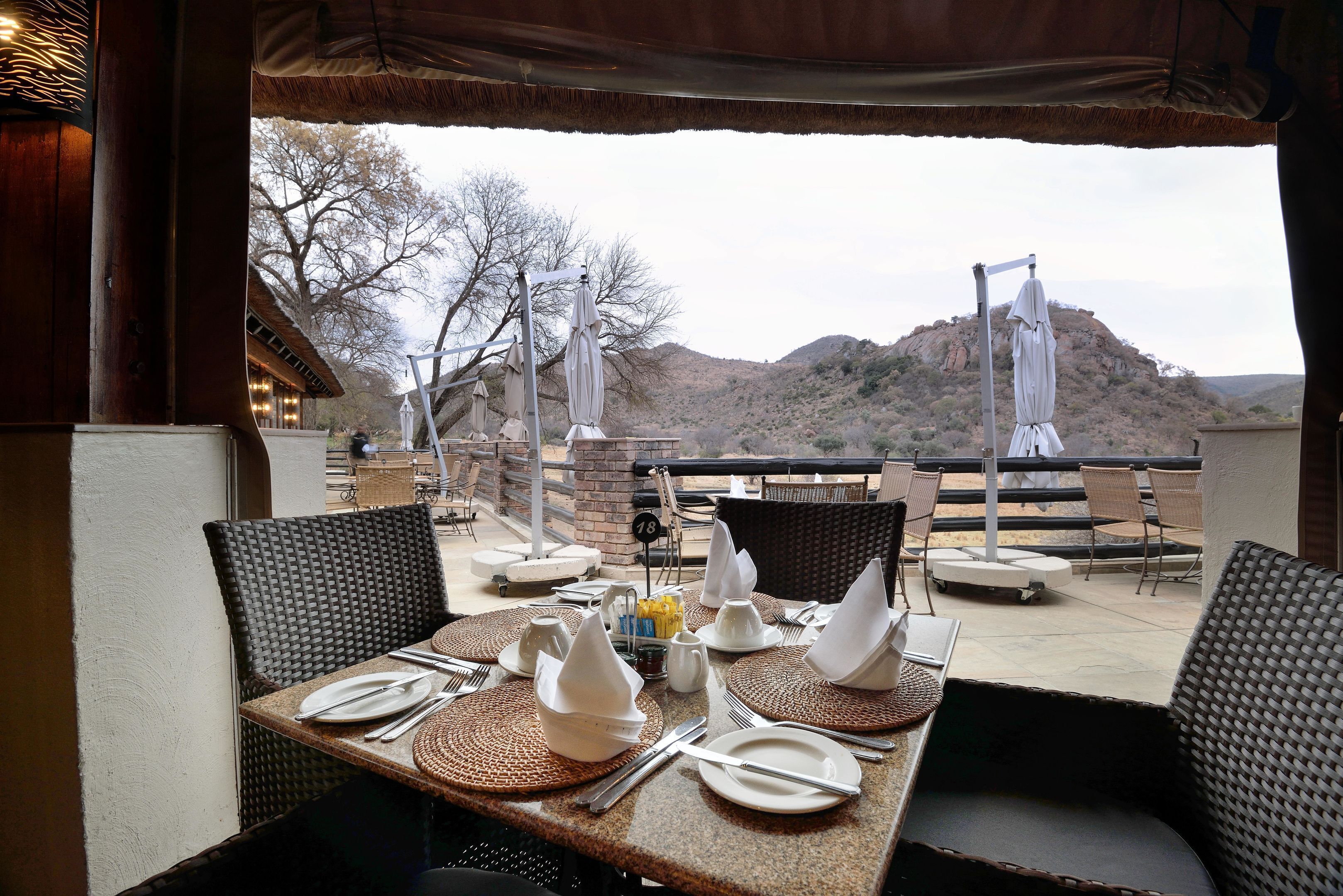 An outdoor dining area with tables set for two, equipped with white plates, silverware, and napkins. The setting includes wicker chairs and a view of a mountainous landscape.