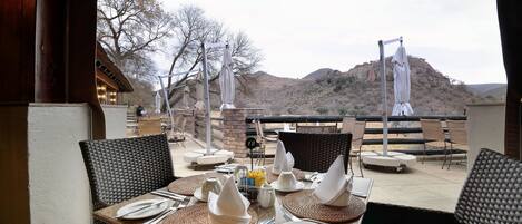 An outdoor dining area with tables set for two, equipped with white plates, silverware, and napkins. The setting includes wicker chairs and a view of a mountainous landscape.