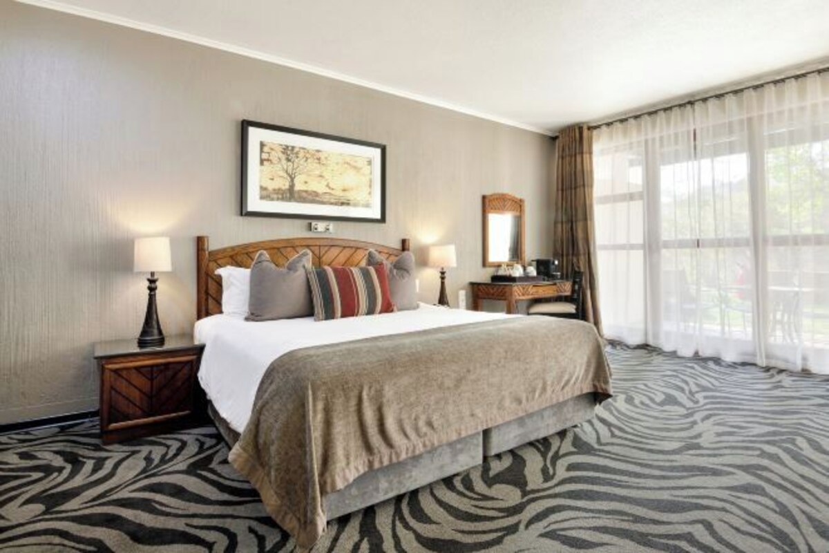 Habitación de hotel con una cama grande, una mesita de noche, una lámpara, un espejo y una ventana con cortinas.