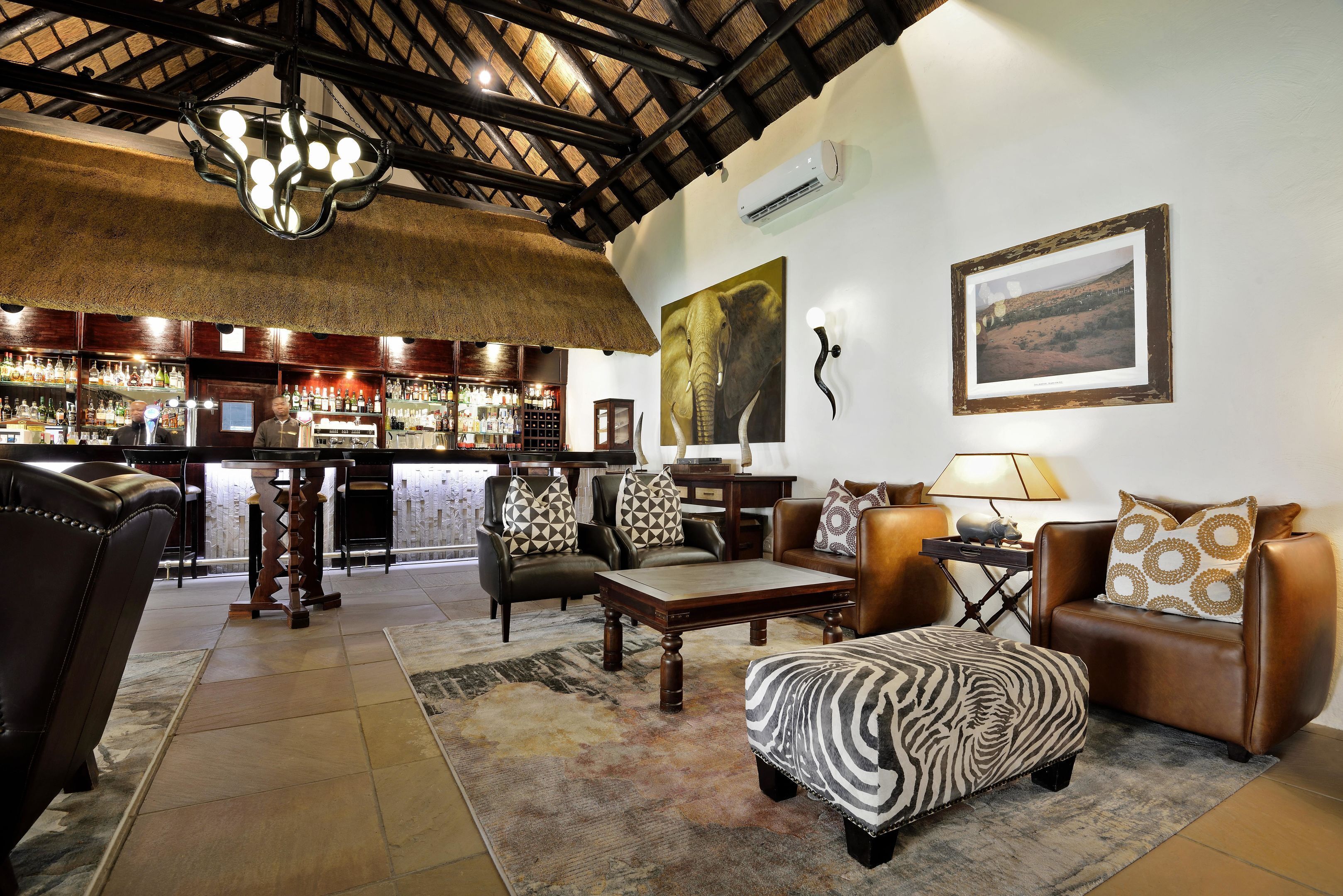 A bar area with wooden furniture and a thatched ceiling, a seating area with leather sofas and patterned cushions, and a large framed picture on the wall.