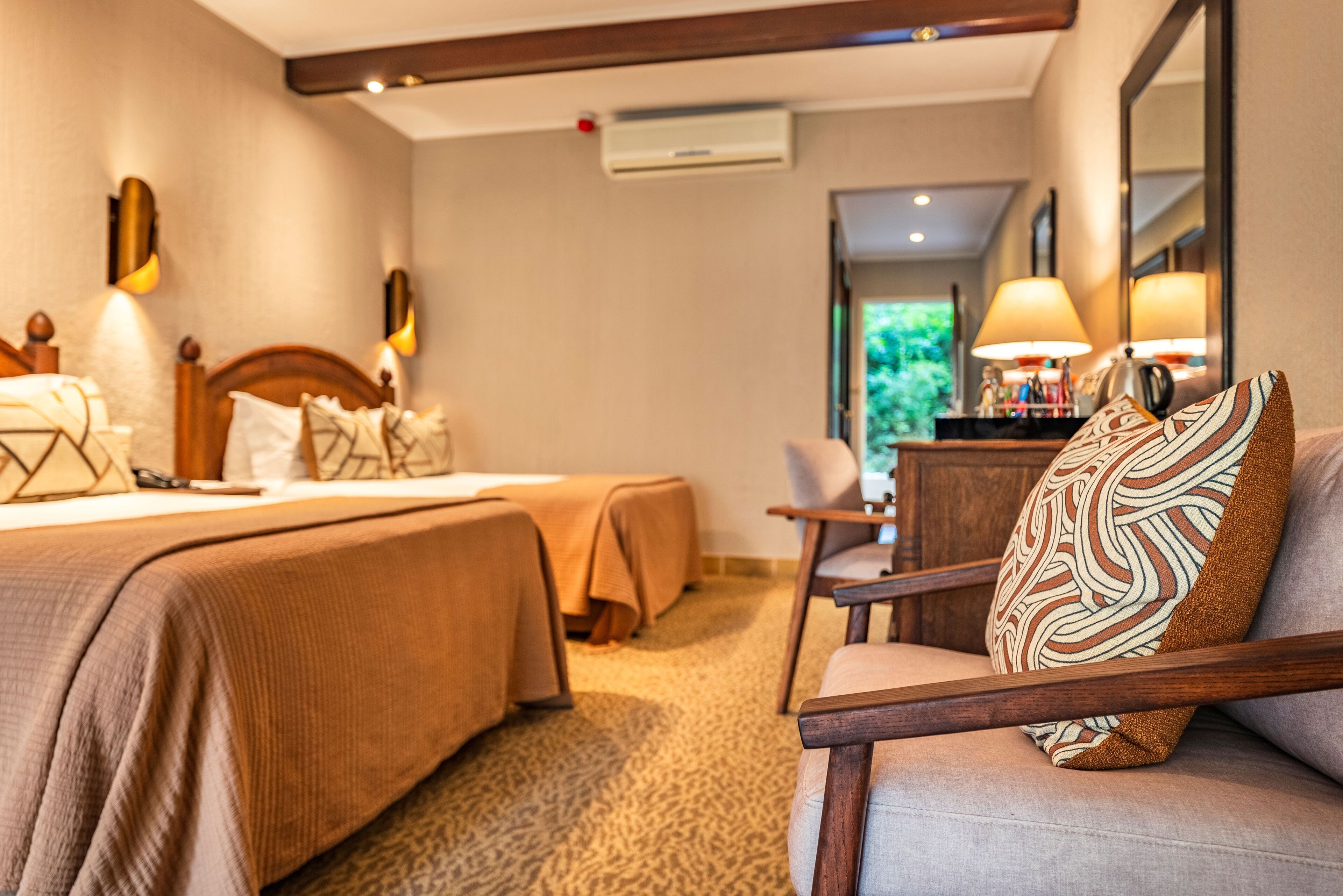 A hotel room with two beds, a sitting area with a chair, and a view of greenery outside.