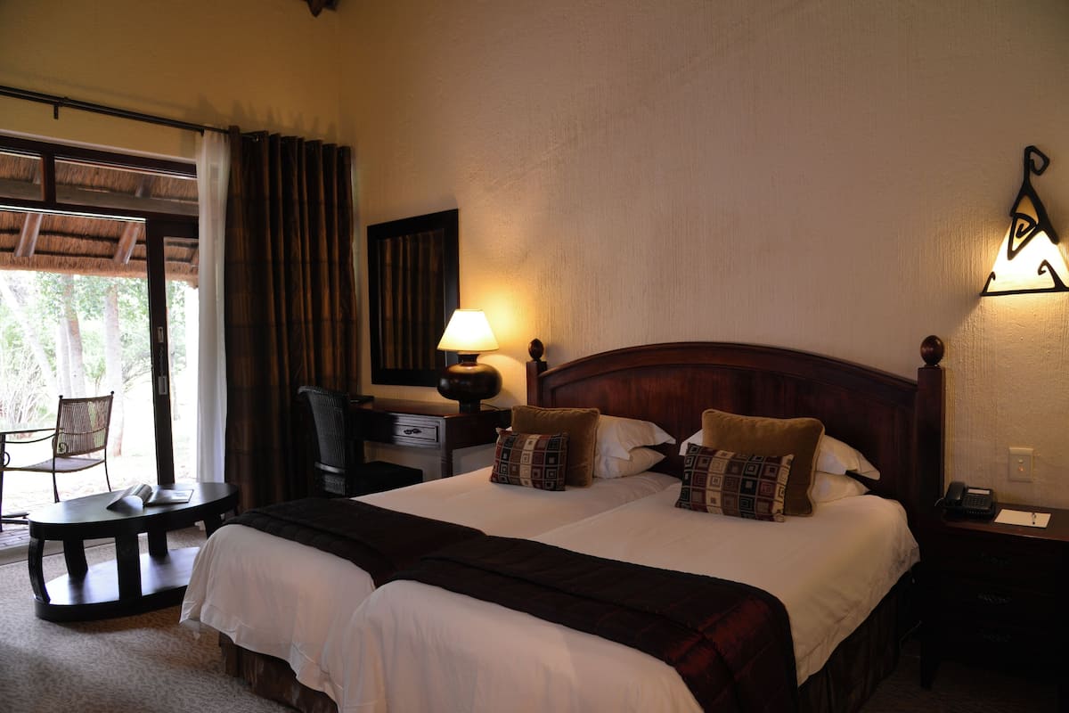A double bed with two pillows, a bedside table with a lamp, a chair, and a desk with a telephone.
