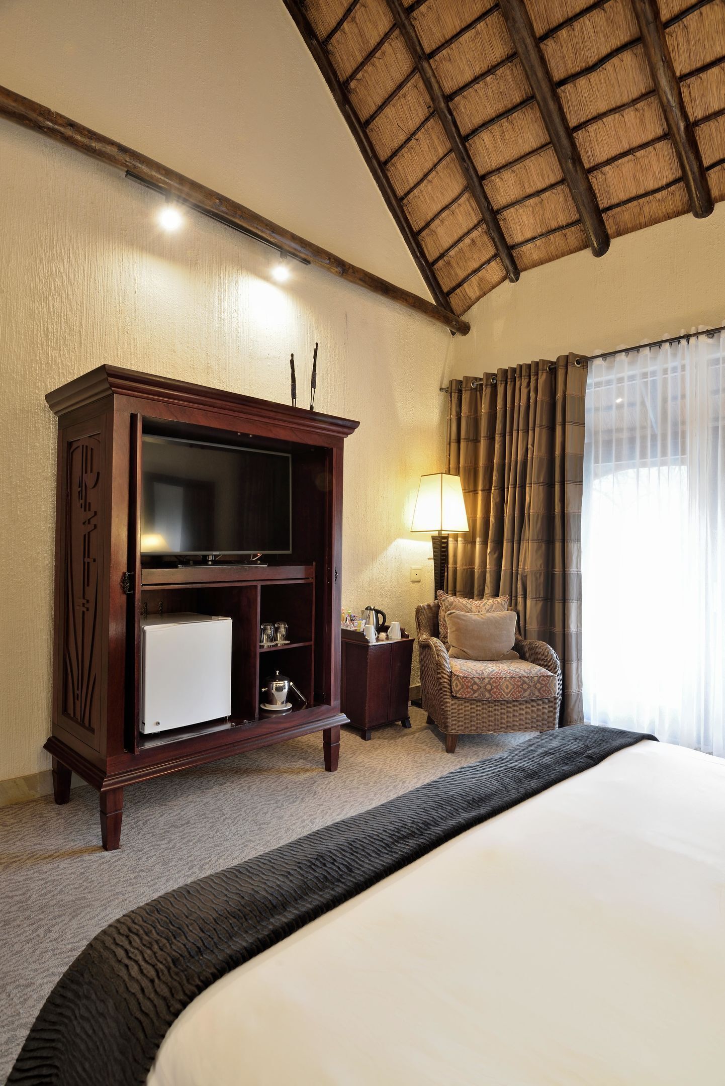 A hotel room with a large bed, a wooden TV stand, a chair, a lamp, and a window with curtains.