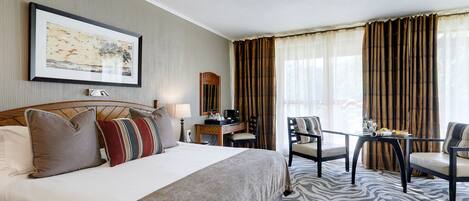 A hotel room with a large bed, a desk, two chairs, a table with a tray of items, and a framed picture on the wall.
