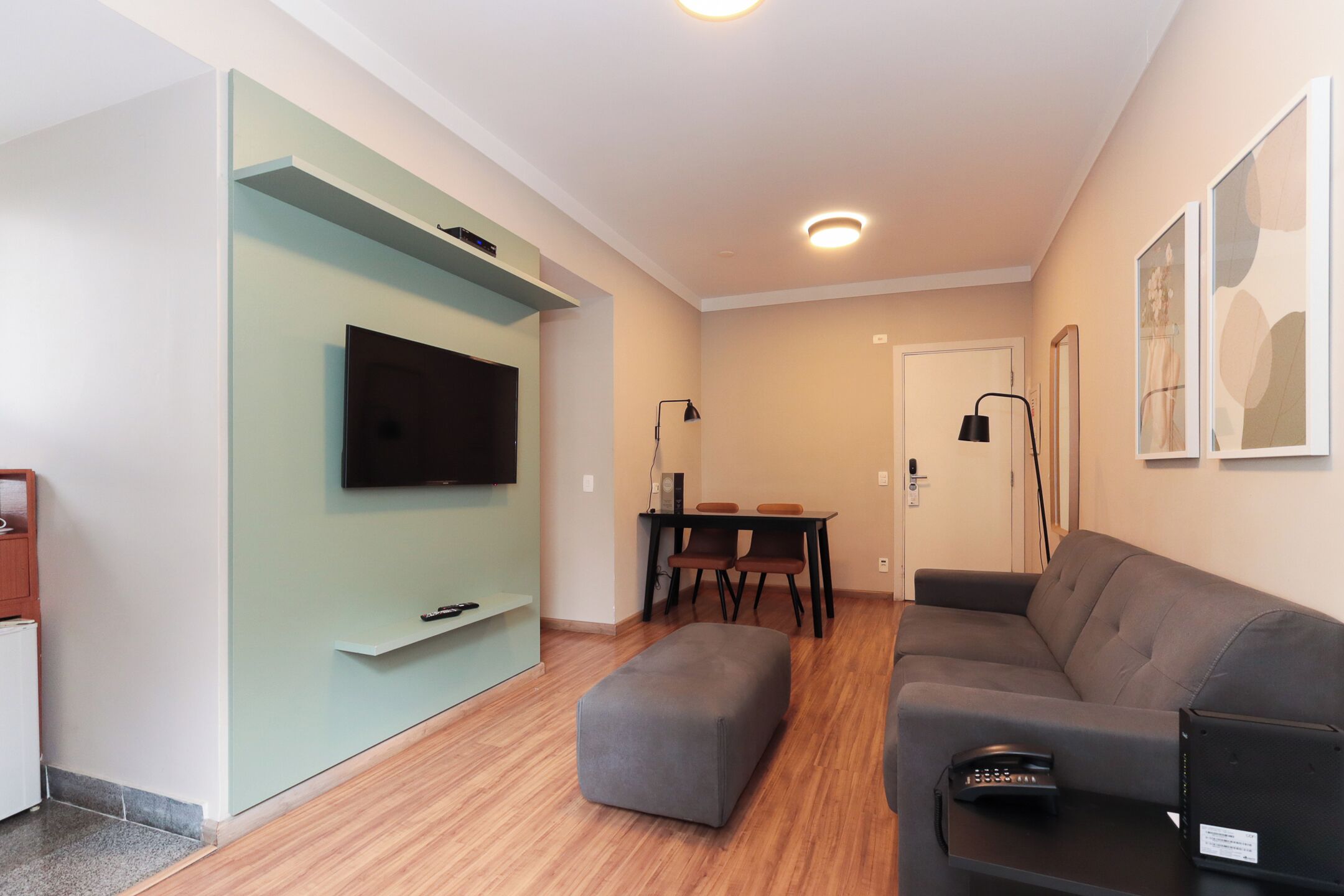 A modern living room with a flat-screen TV, a grey ottoman, a sofa, a dining table with chairs, and a floor lamp.