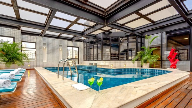 A modern indoor swimming pool with a wooden deck, tiled walls, and large windows. - H4 Hotel Opera Jardins (São Paulo)
