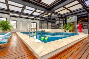 Piscina interior moderna com deck de madeira, paredes revestidas e janelas amplas.