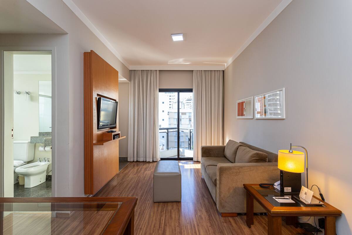 Habitación de hotel con suelo de madera, un sofá, un televisor, una lámpara y un balcón con cortinas.