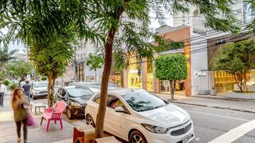 A street scene with a white car parked on the side, a pink chair, and a tree in the foreground.