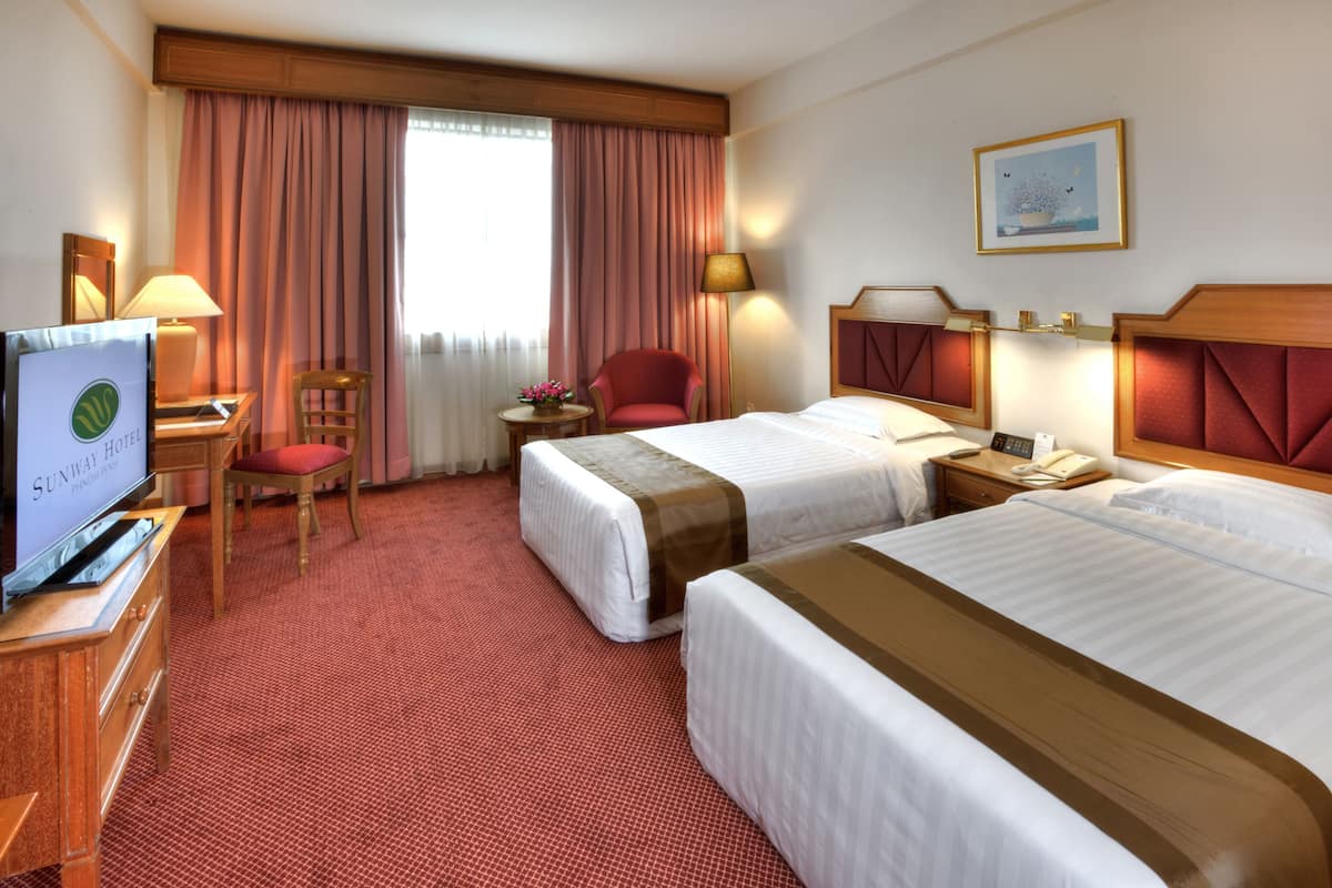 Una habitación de hotel con dos camas, un escritorio con televisor, una silla, una mesita de noche con lámpara y una ventana con cortinas.