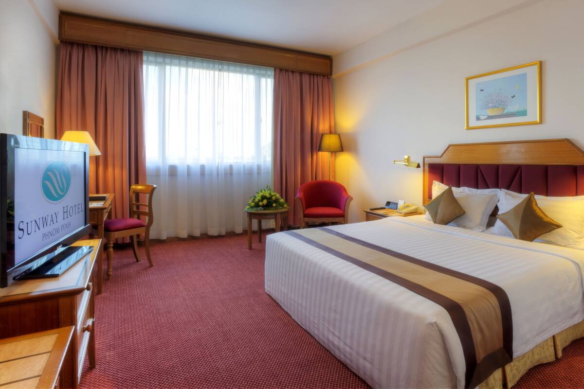 Una habitación de hotel con una cama grande, un escritorio con televisor, una silla roja, una pequeña mesa con flores y una lámpara.