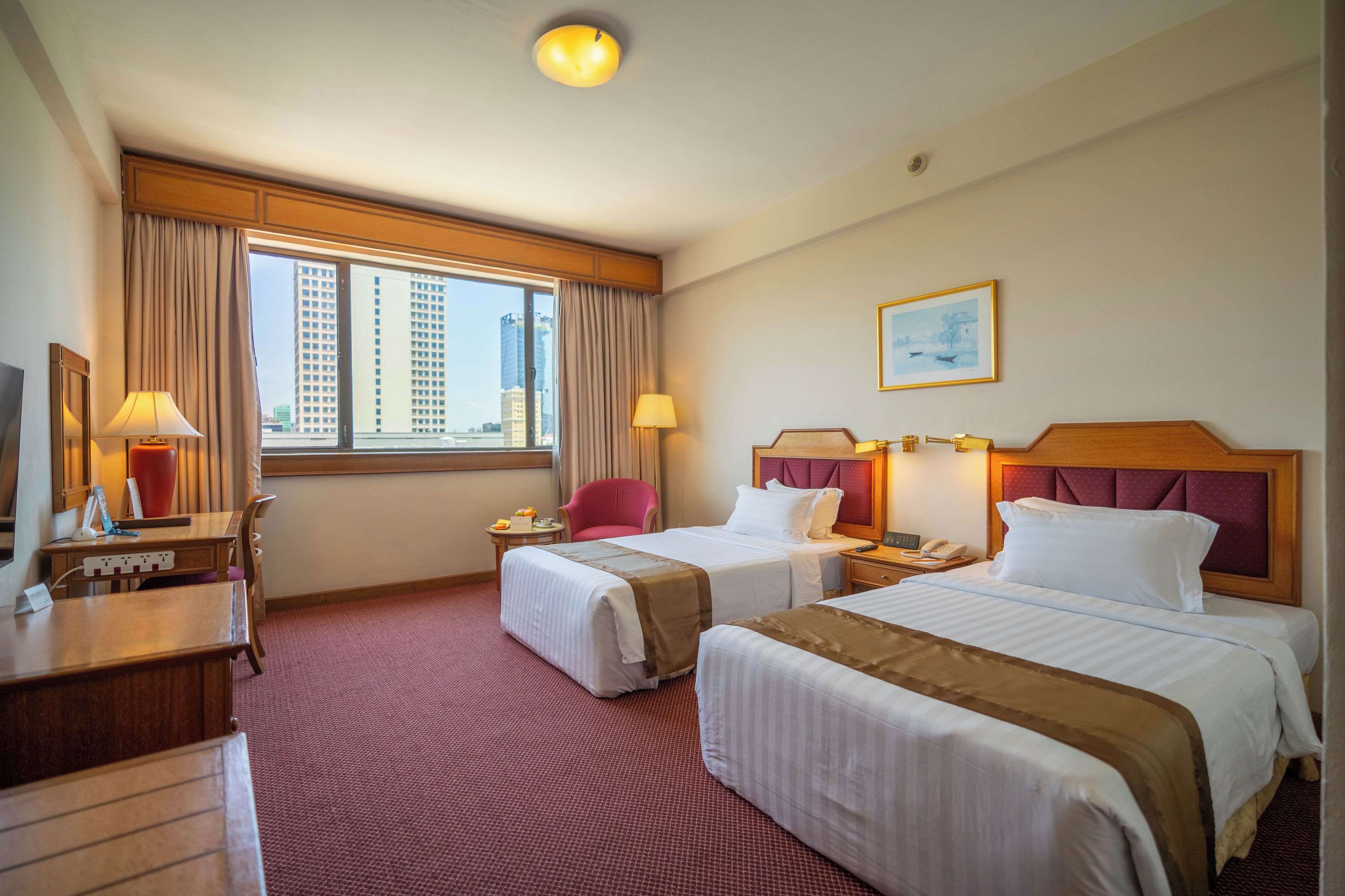 A hotel room with two beds, a desk, a chair, a window with curtains, a painting on the wall, and a ceiling light.