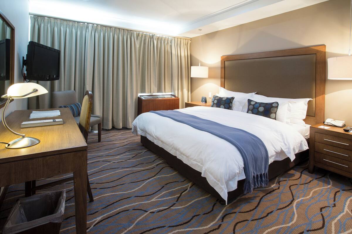 Ein Hotelzimmer mit einem großen Bett, einem Schreibtisch mit Lampe, einem Sessel, einem Fernseher und einem Nachttisch mit Telefon.