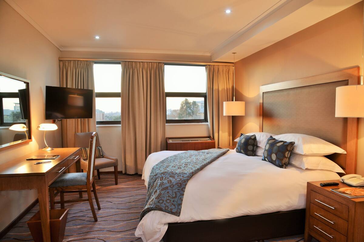 Ein Hotelzimmer mit Bett, Schreibtisch, Stuhl, Fernseher und fensterbepflanzten Fenstern.