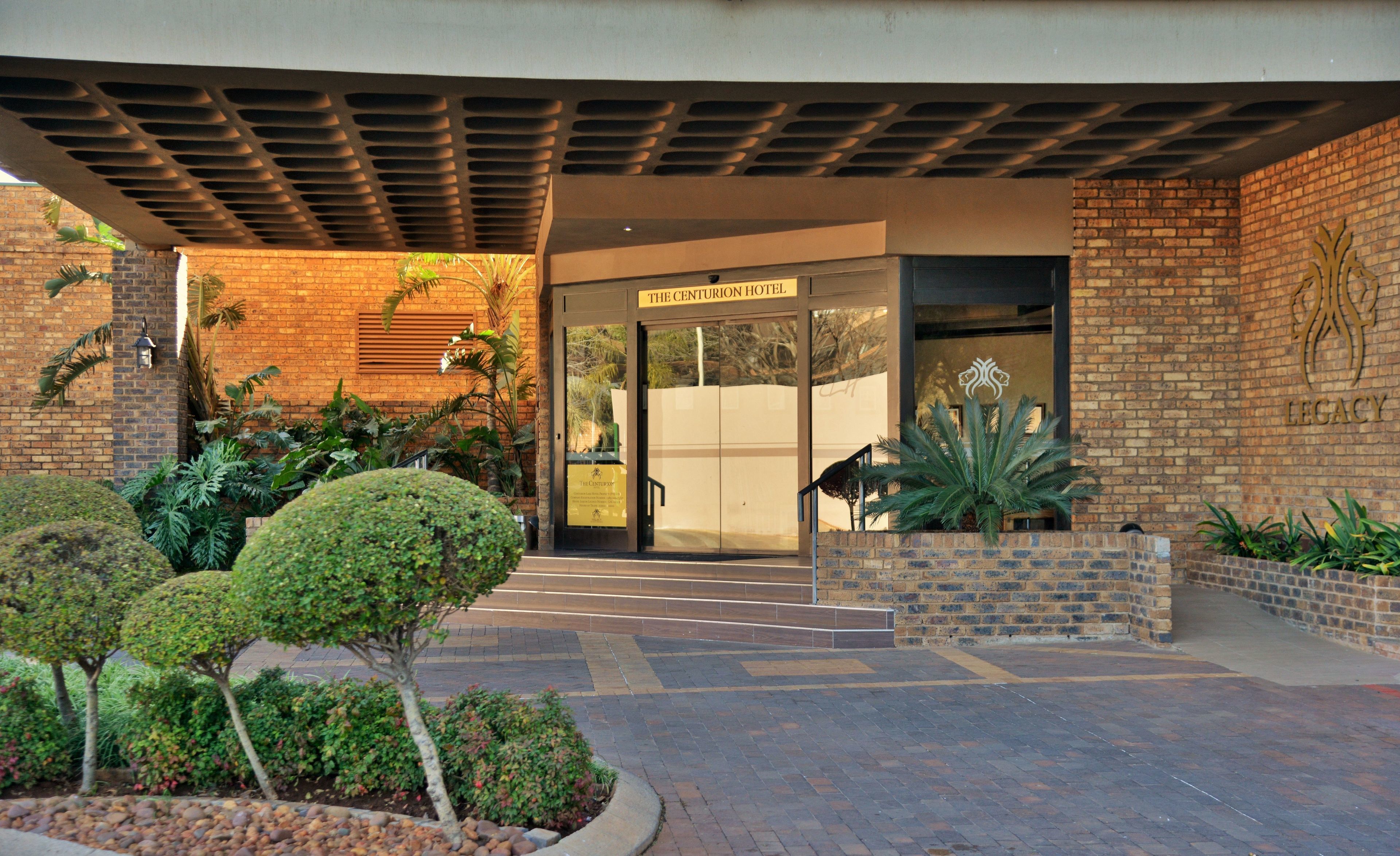 La entrada al Hotel Centurion, con fachada de ladrillo, puerta de vidrio y un jardín ajardinado con arbustos y una palmera.