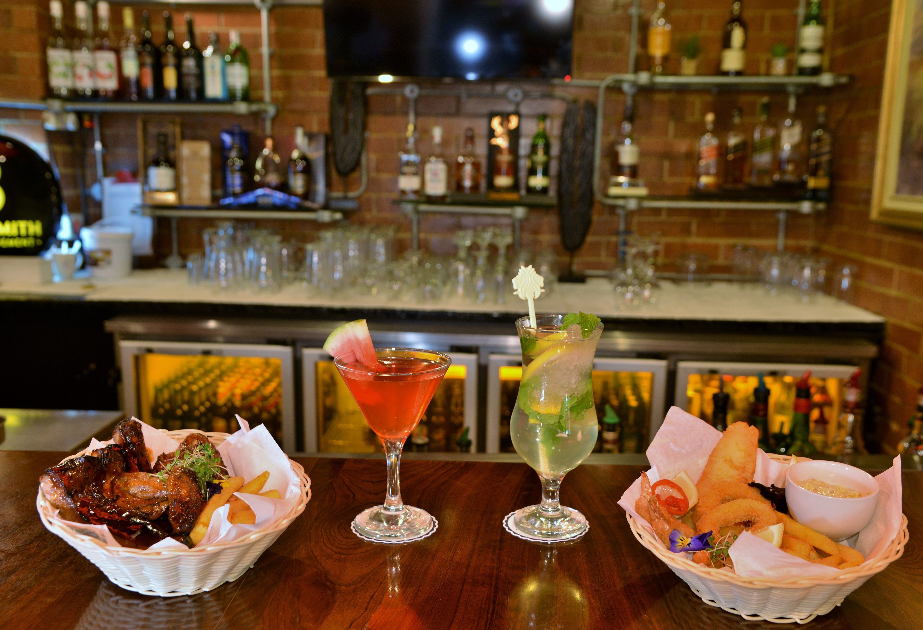 Uma área de bar com três cestos de comida, dois coquetéis e uma variedade de garrafas ao fundo.