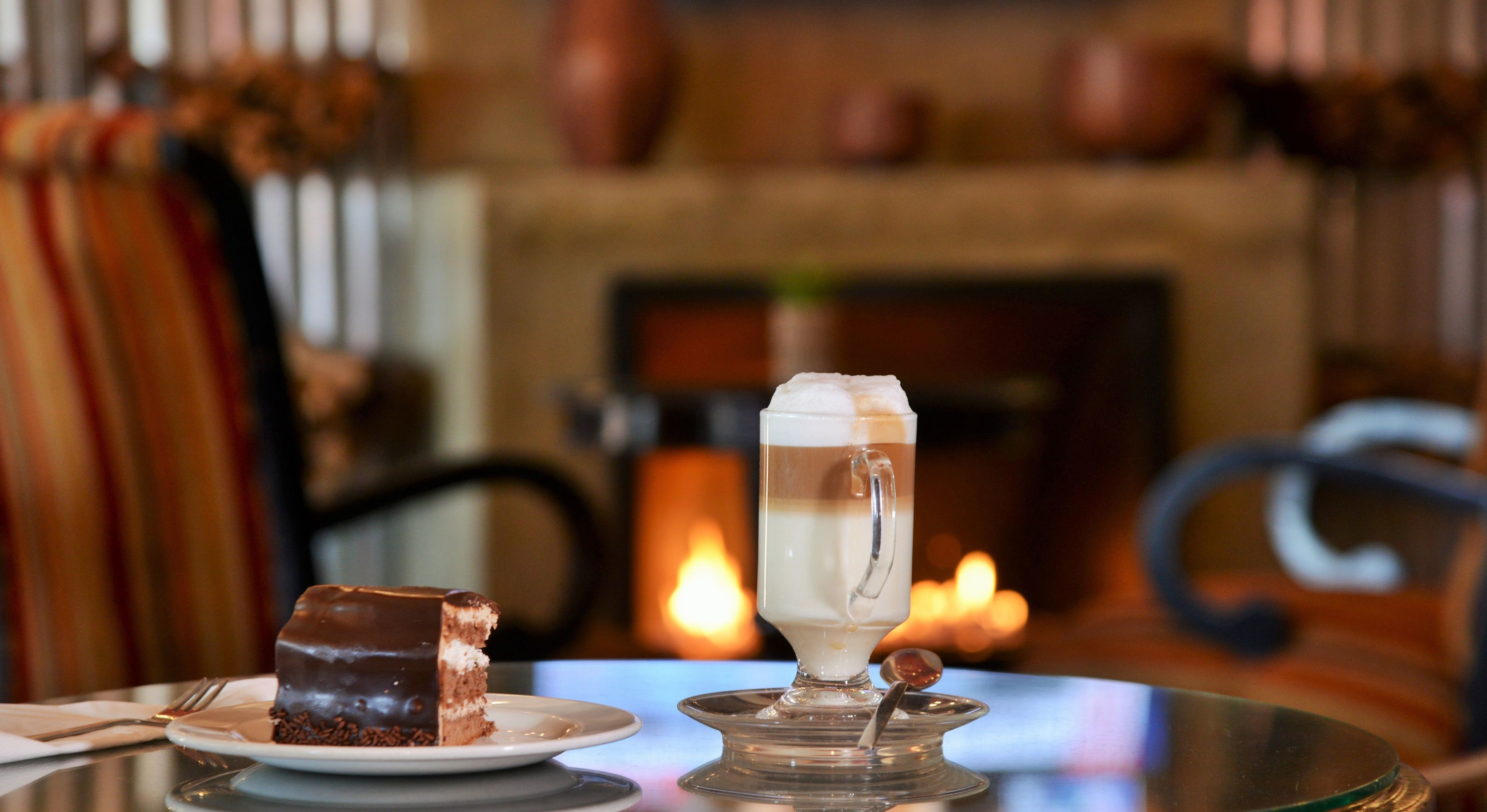 Ein Desserttisch mit einem Kuchenstück und einem Glas mehrschichtigem Getränk, vor verschwommenem Hintergrund mit Kamin.