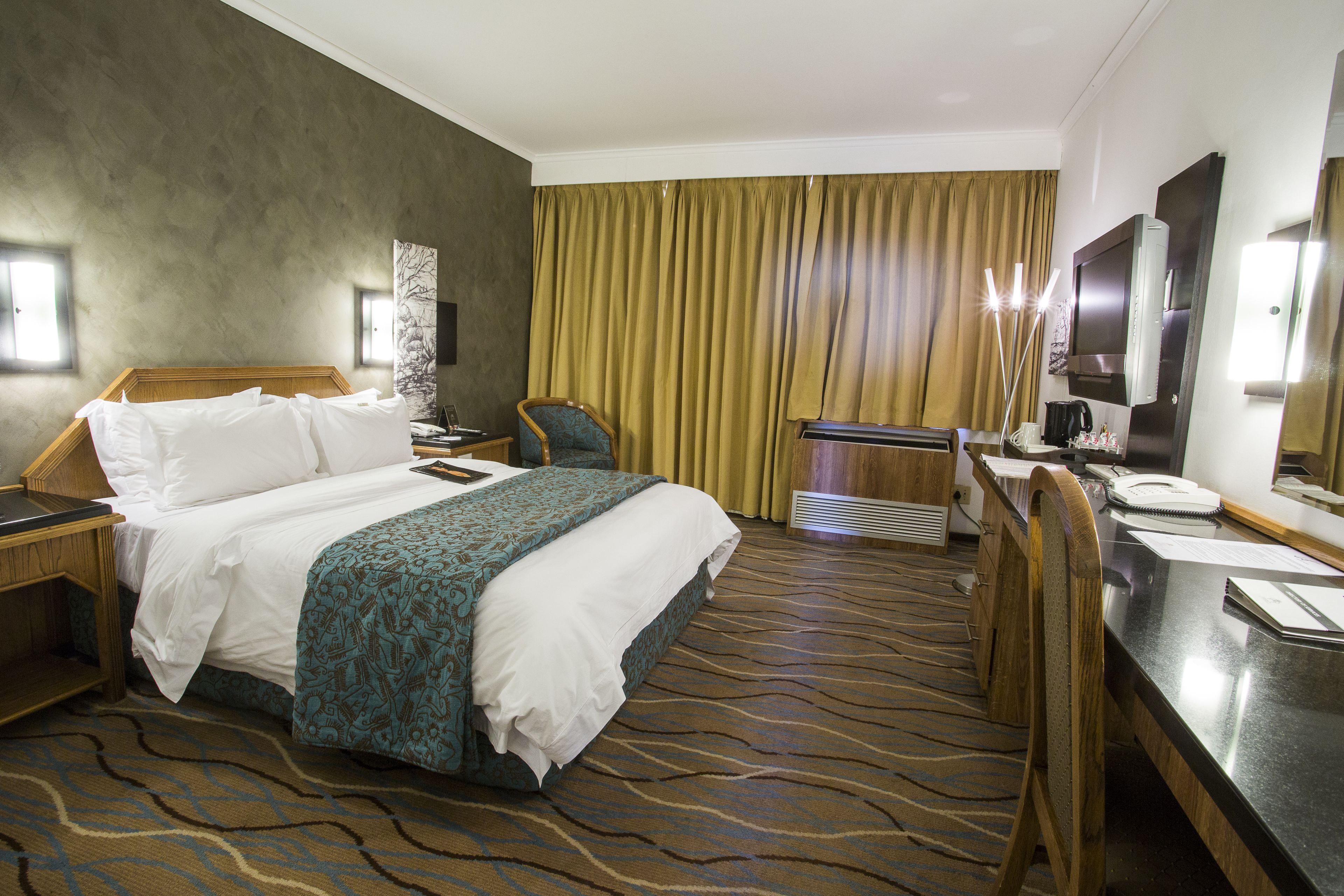 Habitación de hotel con cama, escritorio, silla, televisor y un amplio ventanal con cortinas.