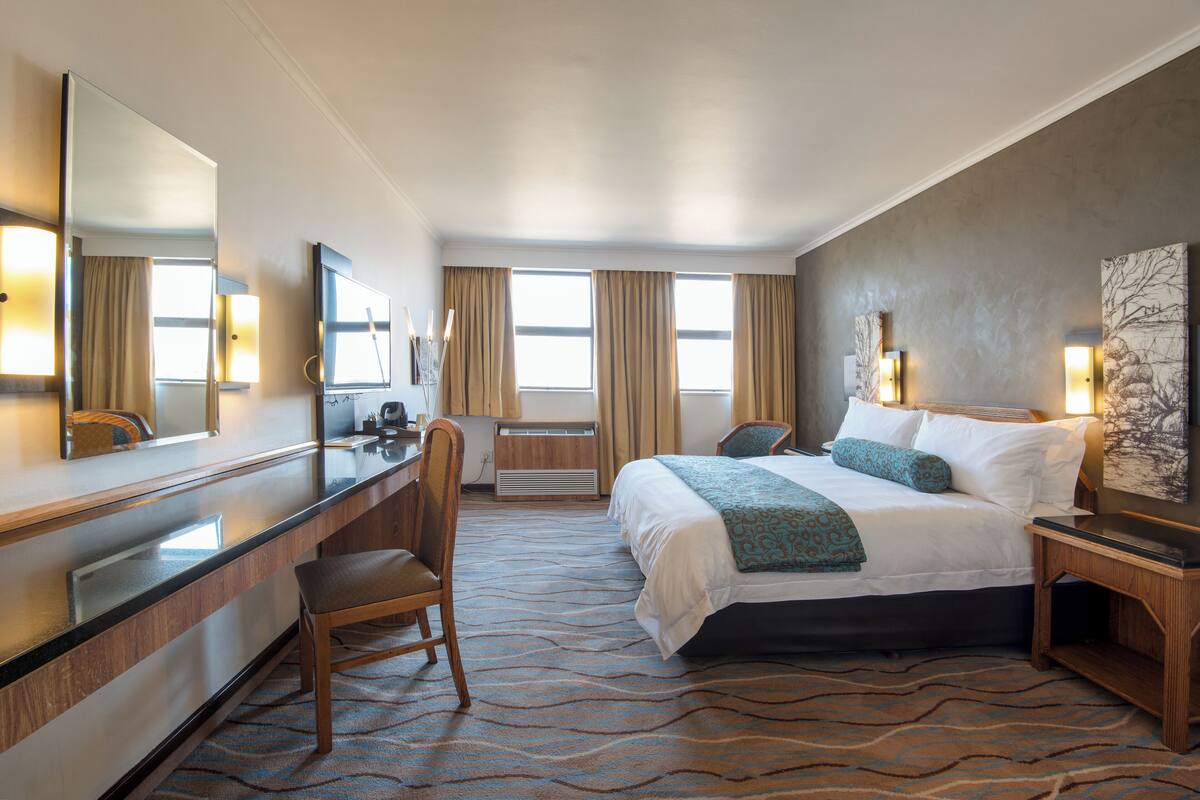 Ein Hotelzimmer mit Bett, Schreibtisch, Stuhl, einem großen Spiegel und einem Fenster mit Vorhängen.