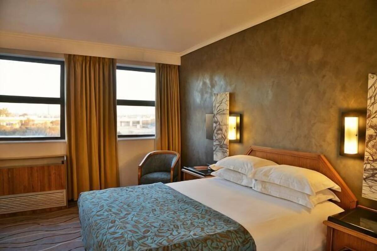 Ein Hotelzimmer mit einem großen Bett, einem Sessel, einem Nachttisch und Blick auf die Stadt durch große Fenster.