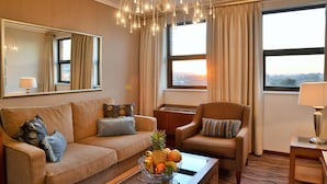 Habitación de hotel con sofá, sillón y una mesa de centro con fruta. Cuenta con un ventanal con cortinas, un candelabro y un espejo en la pared.