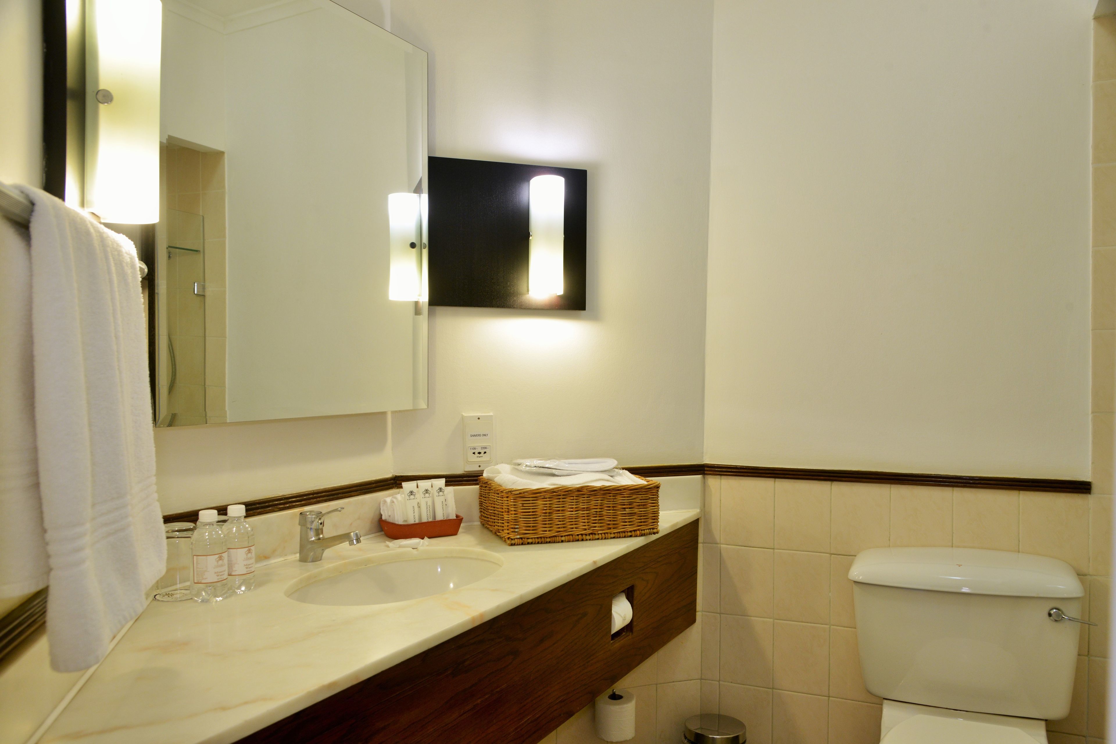 A bathroom with a large mirror, two sinks, a toilet, and a basket of toiletries.