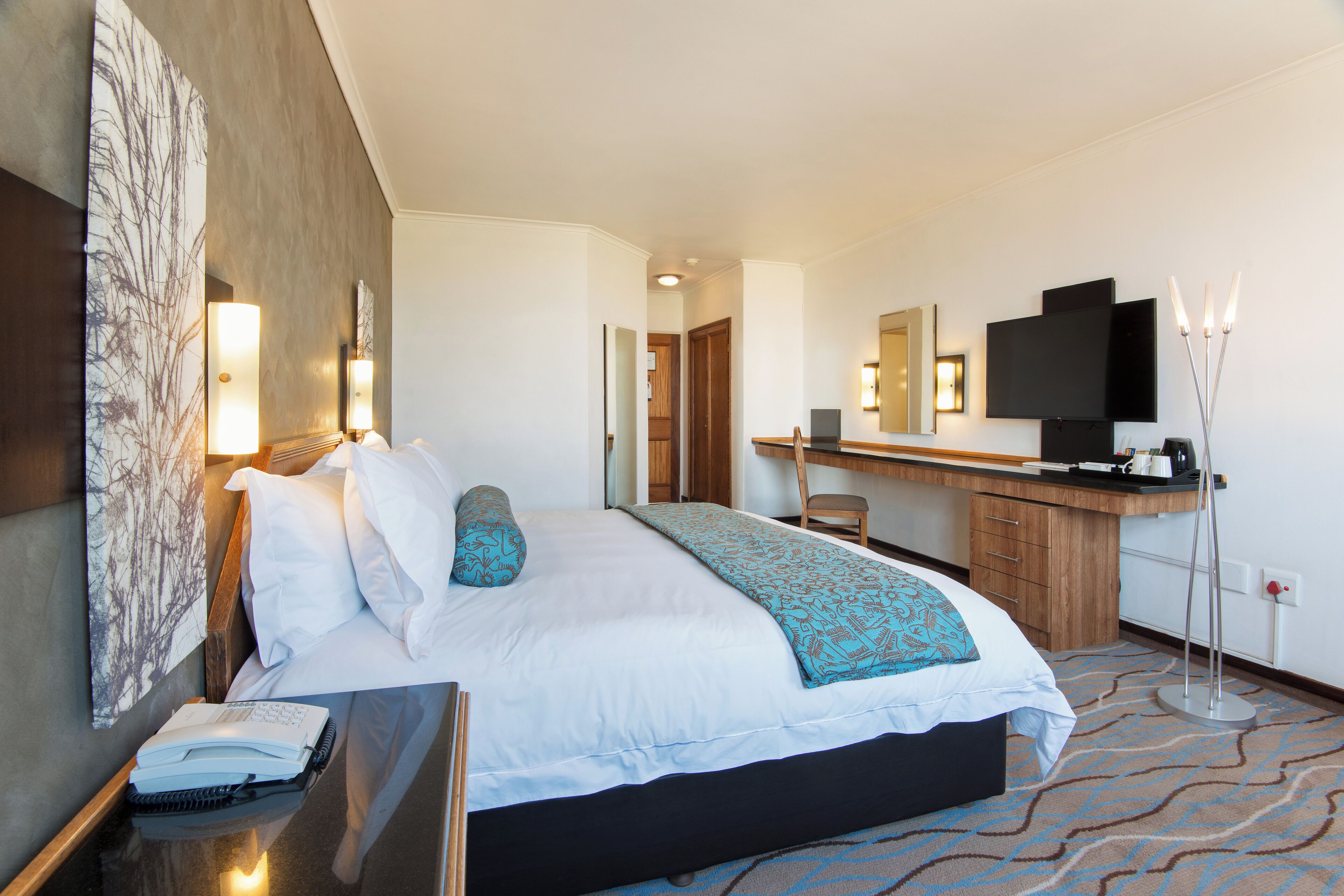 Quarto de hotel com cama, mesa de cabeceira, escrivaninha e televisão.