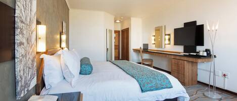 Quarto de hotel com cama, mesa de cabeceira, escrivaninha e televisão.