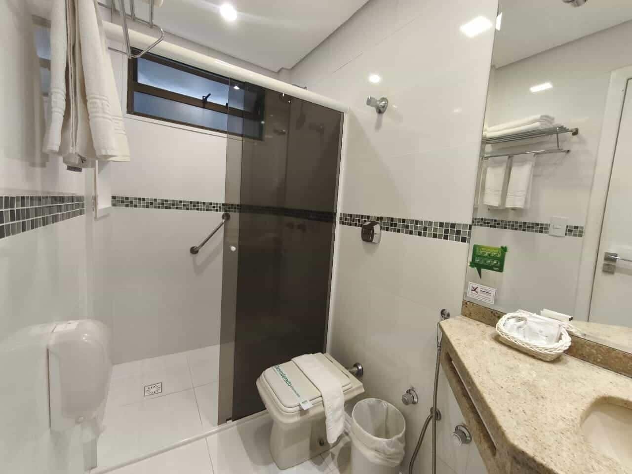 A modern bathroom with a glass shower enclosure, a toilet with a storage bin, a sink with a towel rack, and a mirror with a towel hanging on it.