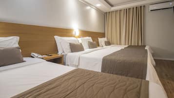 Quarto de hotel com três camas, cabeceira de madeira, mesa de cabeceira com telefone e controle remoto, e ar-condicionado.