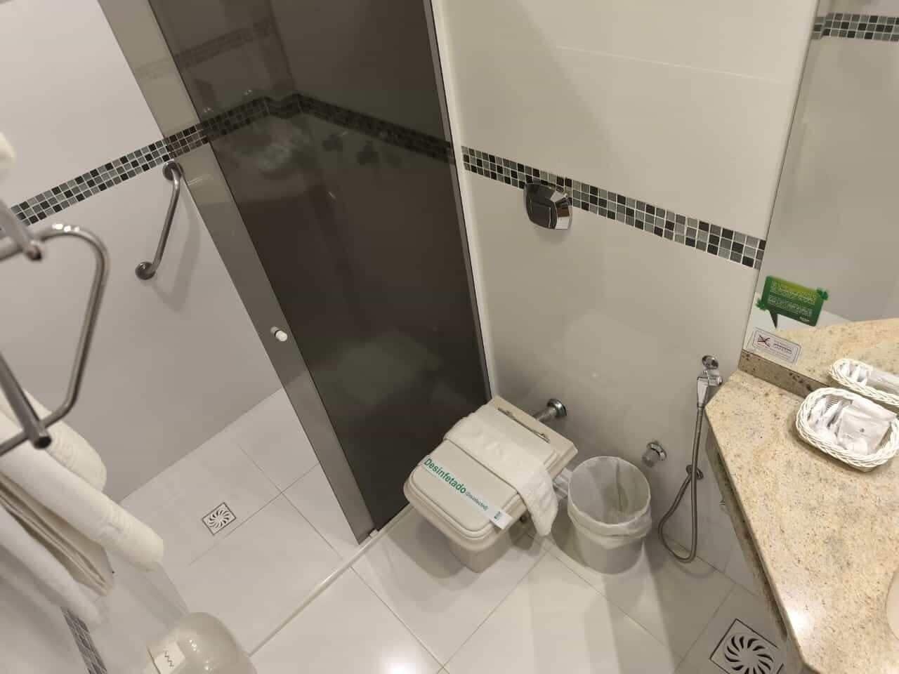 Casa de banho com cabine de duche em vidro, sanita com dispensador de papel, lavatório com saboneteira e um contentor de lixo.
