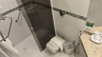 Casa de banho com cabine de duche em vidro, sanita com dispensador de papel, lavatório com saboneteira e um contentor de lixo.